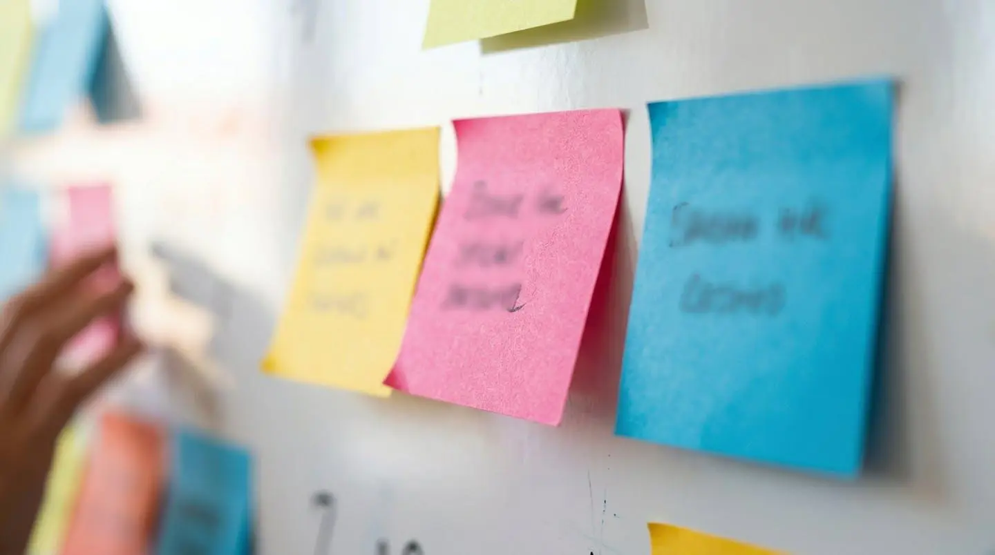 Gros plan sur des post-it colorés collés sur un tableau blanc avec des mains floues en arrière-plan ajoutant des notes